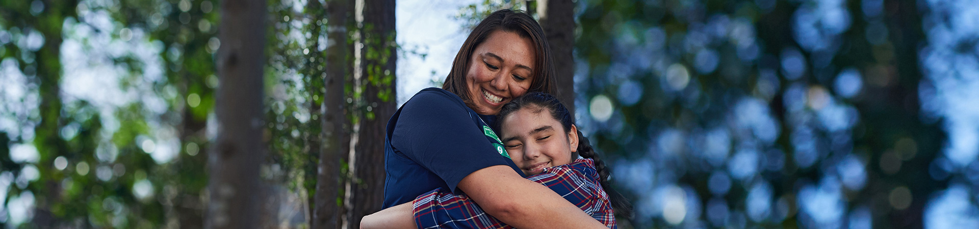  smiling adult volunteer in uniform hugging a Girl Scout outside on a sunny day in the woods 