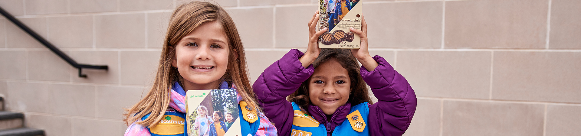 Girl Scout Daisies standing outside holding boxes of cookies and smiling 